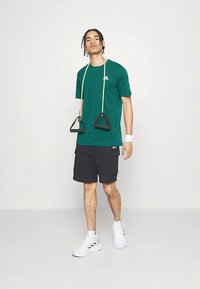 Tânăr îmbrăcat cu un tricou verde Adidas și pantaloni scurți negri ținând o bandă de rezistență, stând pe un fundal simplu deschis.