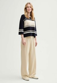 Pull rayé marine et crème avec des manches trois-quarts, associé à un pantalon beige ample et des chaussures beiges, sur un fond neutre.