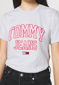 Šedé bavlněné tričko s výrazným červeným nápisem "TOMMY JEANS" a malou akcentací loga Tommy Hilfiger na spodním okraji. Krátké rukávy, klasický střih.