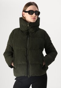 Jachetă din corduroy de culoare verde măslinie, cu guler înalt, închidere cu fermoar și buzunare frontale, având o textură ribbed și o croială lejeră.