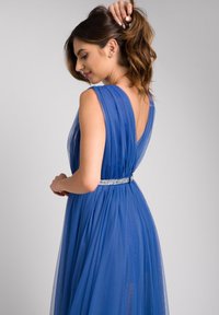 Niebieska sukienka z przezroczystym materiałem, plisowanym fasonem, głębokim dekoltem na plecach i wyszywaną taśmą w pasie. Gładka struktura i swobodny krój.