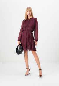 Woman wearing a long-sleeve burgundy buttoned dress with a waist tie, holding a black handbag and wearing black high-heeled sandals.