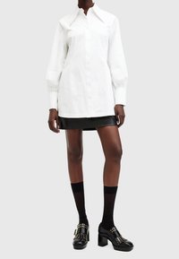 Femme portant une robe chemise blanche à manches longues avec une mini jupe en cuir noire, des chaussettes noires mi-mollet et des chaussures à talons cloutées noires.