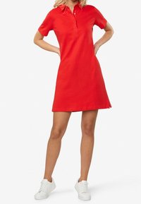 Robe polo rouge à manches courtes, col boutonné et ourlet droit, en tissu de coton lisse. Portée avec des baskets blanches.