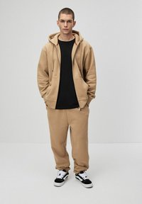 Beżowa bluza z kapturem na zamek z przodu i dużymi kieszeniami, w zestawie z dopasowanymi beżowymi spodniami dresowymi. Czarna koszulka pod spodem, czarno-białe sneakersy.