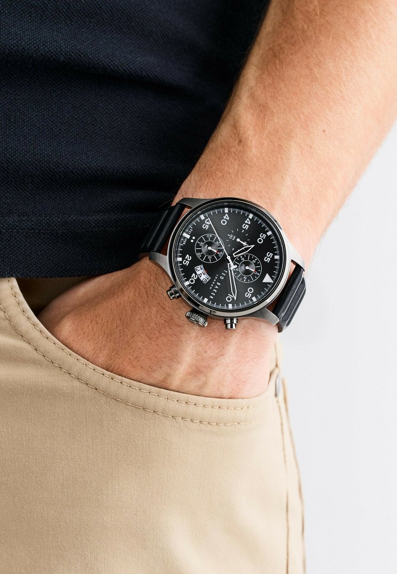 Orologio cronografo nero con cassa in metallo, cinturino nero testurizzato, indicatori delle ore bianchi e sub-quadranti. Indossato su un polso contro un tessuto beige.