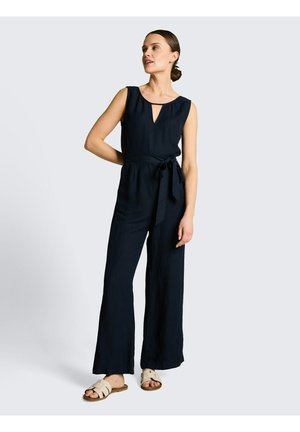 Frau trägt ärmellosen schwarzen Jumpsuit mit gebundener Taille und beigen Sandalen, steht vor hellem grauem Hintergrund.