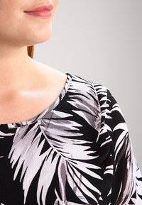 Robe noire et blanche avec un motif de feuilles audacieuses, tissu texturé et un col arrondi, partiellement visible au niveau de l'épaule et du cou.