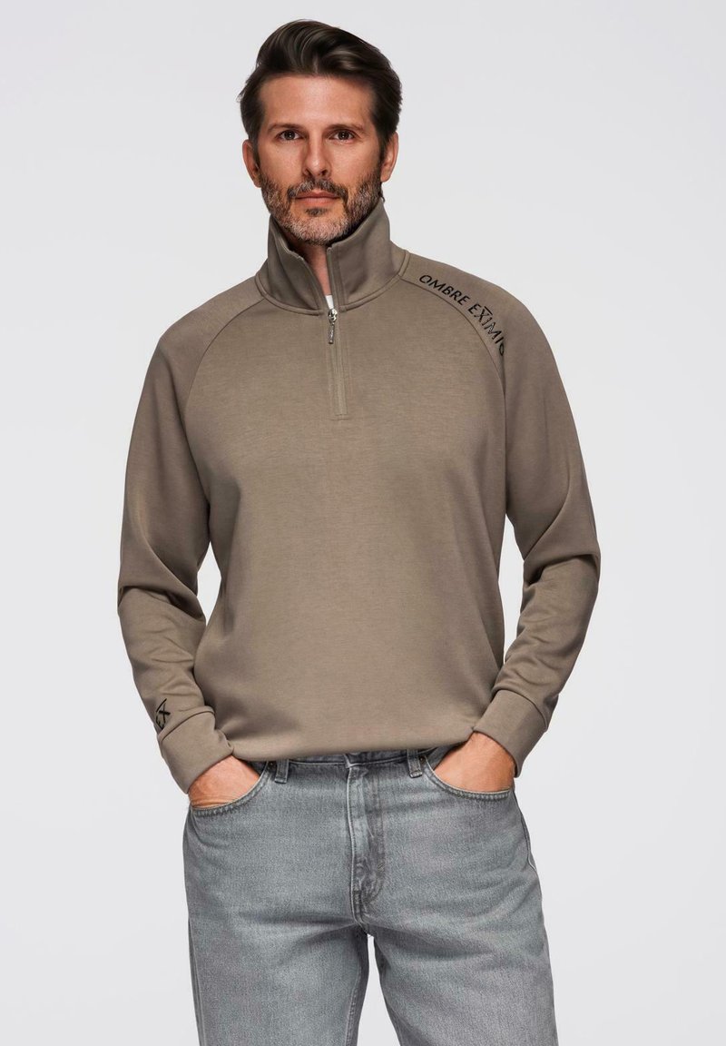 Sudadera de cremallera en color marrón claro con mangas largas y diseño raglán. Presenta un sutil logotipo en el hombro y una tela texturizada. Combinada con jeans grises.
