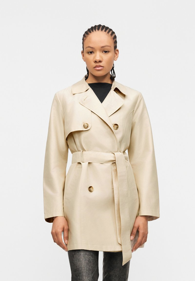 Γυναίκα με πλεξούδες, ντυμένη με μπεζ καμπαρντίνα με ζώνη πάνω από μαύρο τοπ και σκούρο γκρι παντελόνι, στέκεται μπροστά σε απλό λευκό φόντο.