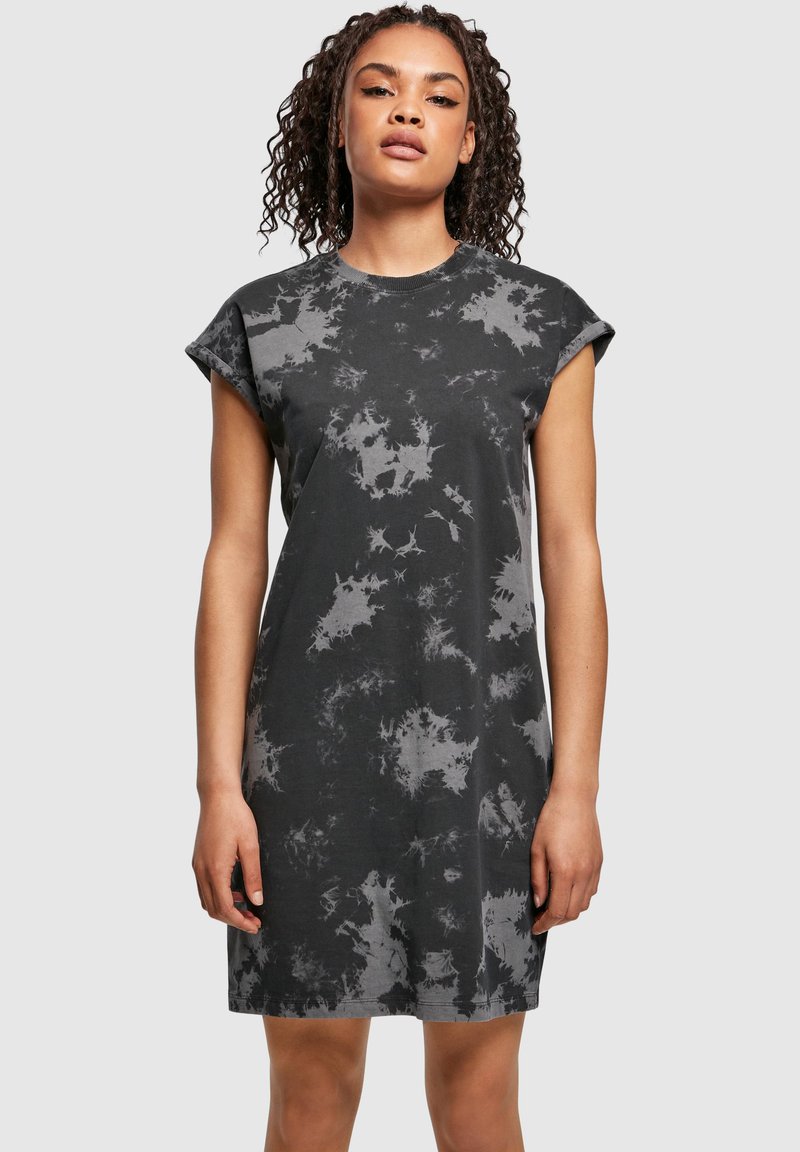 Keeruliste juustega naine, kes kannab musta ja halli tie-dye lühikeste varrukatega t-särgi kleiti, seisab ühetaustalise heleda tausta ees.