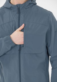 Blå lättviktsjacka med huva, dragkedja fram och ficka. Tillverkad av ett slätt, vattentåligt material med elastiska ärmmuddar.