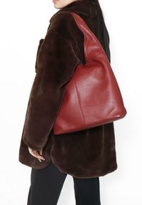 Borsa a spalla in pelle rossa con finitura texturizzata, caratterizzata da un design morbido e rilassato e un fondo arrotondato, indossata sotto un cappotto in pelliccia sintetica marrone.