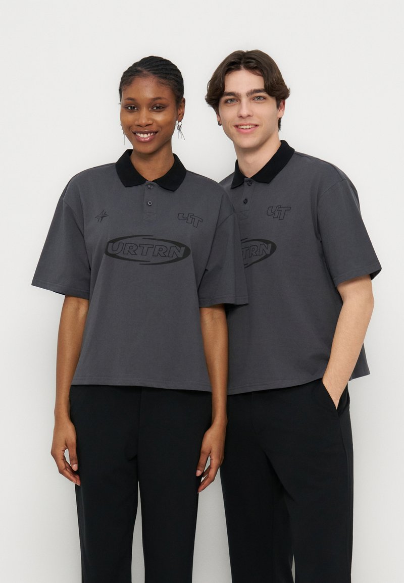 Δύο νέοι ενήλικες, ένας θηλυκός και ένας αρσενικός, φορούν ταυτόσημες σκούρες γκρι πόλο μπλούζες με λογότυπα, στέκονται δίπλα ο ένας στον άλλο μπροστά σε λευκό φόντο.