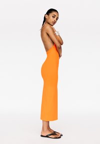 Robe moulante sans manches orange avec une fente haute et un design dos nu, associée à des tongs noires. Texture lisse, silhouette cintrée.