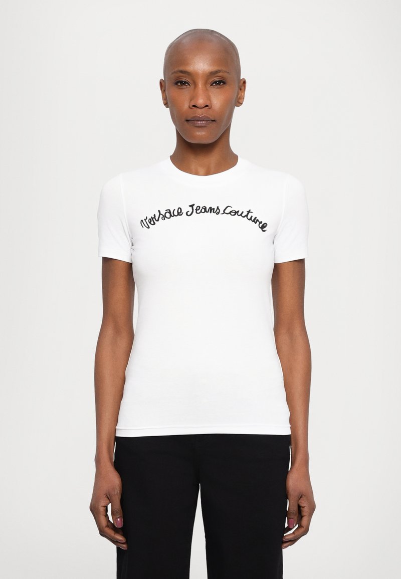 Bílý tričko s krátkým rukávem, přiléhavé, s černým vyšitým textem "Versace Jeans Couture" across hrudníkem. Hladká textilie.