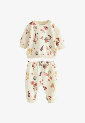 Baby in floralem Outfit sitzt auf einer Decke und hält ein Holzspielzeug in einem sanft beleuchteten Raum mit Holzmöbeln im Hintergrund.