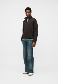 Άνδρας με μαύρο πουλόβερ με φερμουάρ μέχρι το στήθος, πράσινο πουκάμισο από κάτω, μπλε τζιν φαρδιάς γραμμής και λευκά αθλητικά παπούτσια, στέκεται μπροστά σε μονόχρωμο φόντο.