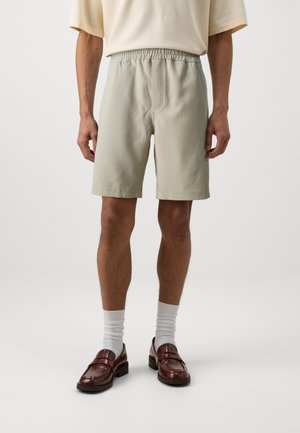 Mand iført beige elastiske shorts med talje, hvide sokker, brune læderloafers og en lys, kortærmet, tekstureret top, stående mod en ensfarvet baggrund.