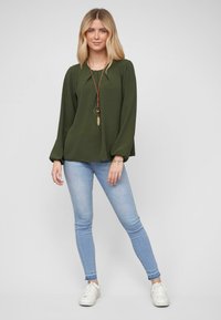Olijfgroene langemouwen top met een ronde hals en elastische manchetten, gecombineerd met lichtblauwe skinny jeans en witte sneakers.