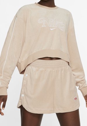 Žena nosící béžovou krátkou mikinu Nike a odpovídající elastické šortky se snapovými knoflíky na bocích.