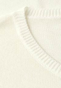 Primo piano di un maglione bianco lavorato a maglia, con un colletto a costine e una lavorazione a trama leggermente allentata. Materiale morbido con leggere sfumature.