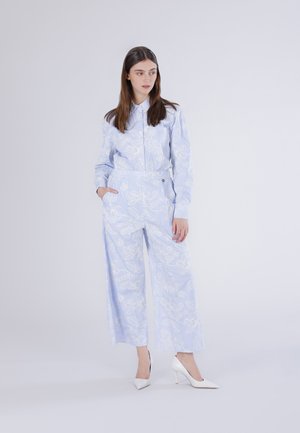 Femme portant une chemise à manches longues bleu clair à fleurs et un pantalon à jambes larges, chaussée de talons hauts blancs, debout avec une main dans la poche et regardant sur le côté.