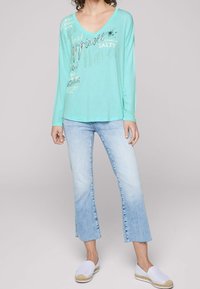 Ljus turkos långärmad tröja med V-halsringning, med silverfärgad grafisk text. I kombination med urblekta blå cropped-jeans och vita slip-on skor.