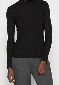 Schwarzes, geripptes Langarm-Turtleneck-Oberteil mit gewelltem Saum und Ärmelabschlüssen, kombiniert mit grauen, strukturierten Hosen. Weiches Material und figurbetontes Design.