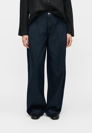Person iført vidde mørkeblå denimjeans, sort skjorte med knapper og sorte sko, stående mod en enkel lys baggrund.