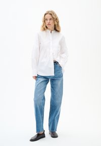 Weißes Hemd mit Knopfleiste und Faltendetail, kombiniert mit hellblauen, lockeren Jeans und schwarzen flachen Schuhen, vor einem einfarbigen Hintergrund.