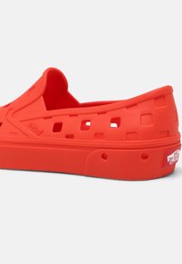 Chaussure rouge en caoutchouc à enfiler avec dessus perforé, bout arrondi et semelle extérieure texturée. Présente le logo Vans sur le talon. Matériau lisse et flexible.