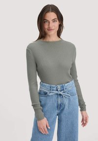 Frau mit glattem braunem Haar, die ein rippendes grau-grünes Langarmoberteil und hoch geschnittene, hellblaue Jeans trägt, die mit einem Jeansband zusammengebunden sind.