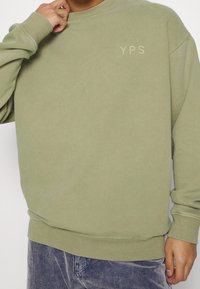 Olive zelená mikina s kruhovým výstřihem, žebrovanými manžetami a lemem. Na hrudi se nachází logo "YPS" ve světlejším odstínu. Měkká textura.