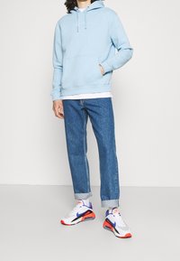 Light blue hoodie with front pocket, worn over a white shirt. Paired with rolled blue jeans and multicolored sneakers.