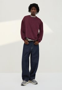 Persoon met korte dreadlocks, gekleed in een losse bordeauxrode trui, wijde donkere spijkerbroek en grijze sneakers, staand tegen een effen lichte achtergrond.