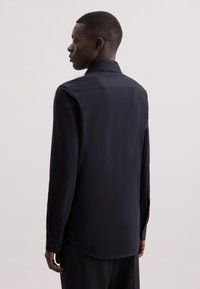 Maglietta nera a maniche lunghe con colletto, texture liscia, orlo curvo e polsini abbottonati, indossata da una persona vista da dietro.