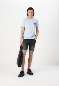 T-shirt en coton bleu clair avec une poche poitrine, associé à un short en denim gris foncé, avec des cuffs retroussés et des chaussures en cuir noires, tenant un sac fourre-tout noir.