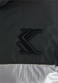 Juodas pūkinis švarkas su iškilusiu juodu siuvinėtu logotipu ant tekstūruotos medžiagos, o apatinėje dalyje yra blizgus sidabrinis finišas.