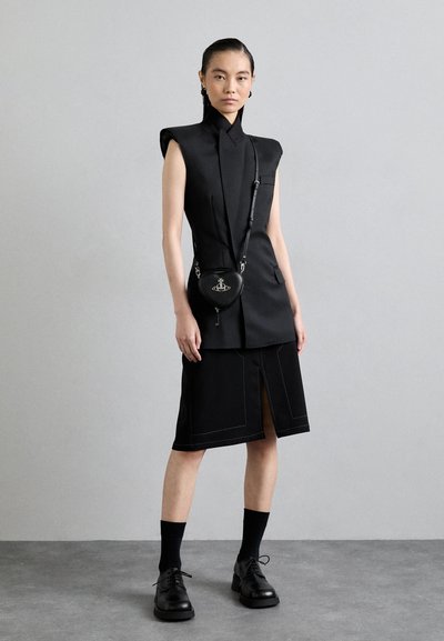 Čierny bezrukávový top s vysokým golierom a priliehavým strihom. V páre s čiernou sukňou po kolená, ktorá má kontrastné prešívanie. Doplnky zahŕňajú malú okrúhlu tašku.