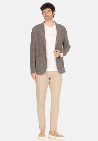 Blazer marrone con texture, due tasche frontali, T-shirt bianca, chinos beige e mocassini color nocciola. Il modello è in posa contro uno sfondo chiaro.
