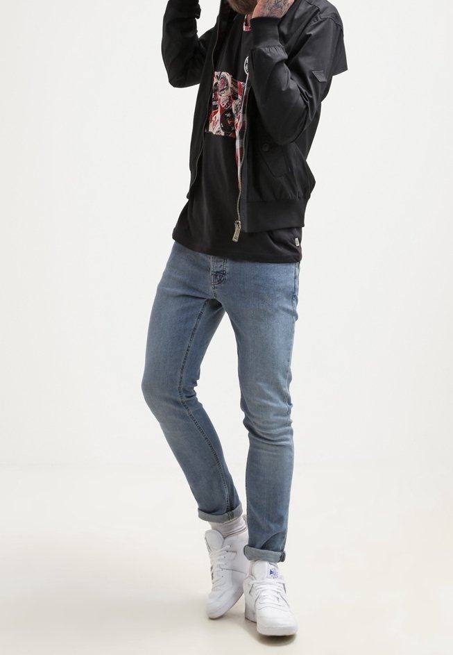 Homme en jean bleu clair, baskets blanches, T-shirt noir à motif, et veste zippée noire, ajustant sa capuche avec les mains près de la tête.