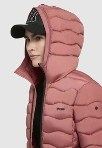 Padded, light pink jacket with a high neck and hood, featuring a black zipper and logo. Black cap with white logo completes the look.