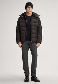 Giacca a vento nera con zip frontale e cappuccio removibile, abbinata a pantaloni grigi scuri a fantasia e stivali neri stringati.