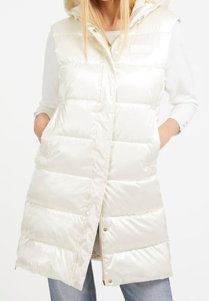 Kobieta ubrana w długą, białą, błyszczącą pikowaną kamizelkę z kapturem, z rękami w kieszeniach, na białej bluzce z długim rękawem i niebieskich dżinsach.