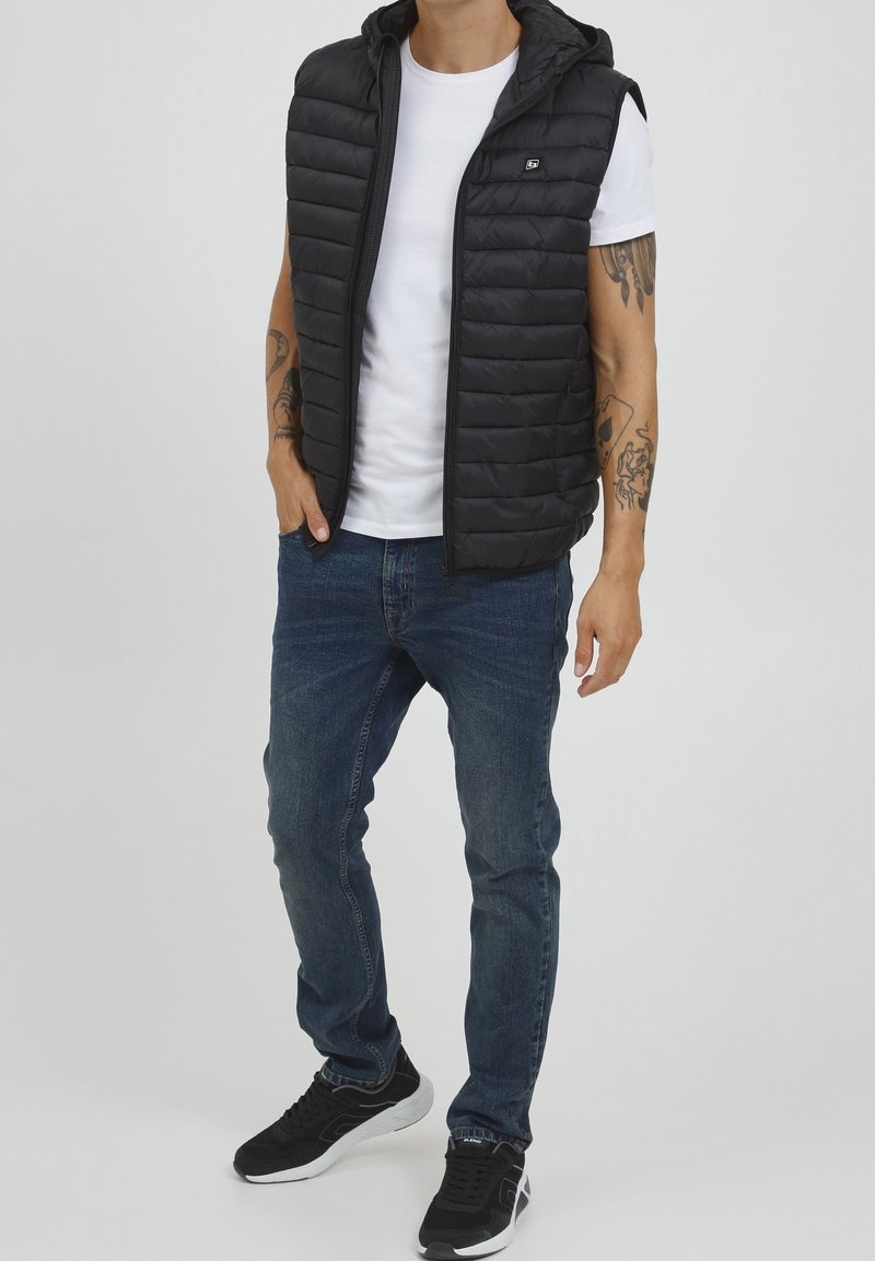 Schwarze gefütterte Weste mit Kapuze, Reißverschluss und kleinem Logo. Getragen über einem weißen T-Shirt, kombiniert mit blauen Jeans und schwarzen Sneakers.