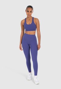 Paarse sportbeha en hoge taille leggings van rekbaar materiaal. Witte sneakers met veters maken de sportieve outfit compleet.
