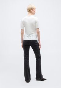 Άτομο με κοντά ξανθά μαλλιά, γυρισμένο πλάτη, φοράει λευκό κοντομάνικο πουκάμισο, μαύρο τζιν και μαύρα παπούτσια με τακούνι.