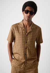 Short-sleeve button-up shirt with brown fabric featuring a repeating beige logo pattern. Sunglasses and simple silver necklace accents.