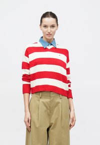 Πλεκτό πουλόβερ με κόκκινες και λευκές ρίγες, με μπλε πουκάμισο με γιακά από κάτω. Το πουλόβερ διαθέτει ριμπ μανσέτες και κοντό στρίφωμα.